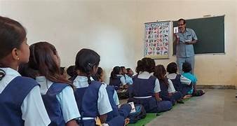 एक सरकारी स्कूल में छात्राएं ज़मीन पर बैठकर शिक्षक से पढ़ाई करते हुए, ग्रामीण भारत की प्राथमिक शिक्षा की जमीनी हकीकत।